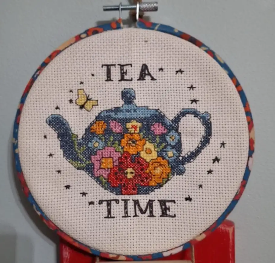 A cross-stitch piece in a circular hoop shows a blue teapot decorated with colorful flowers, with the words 'TEA' above and…