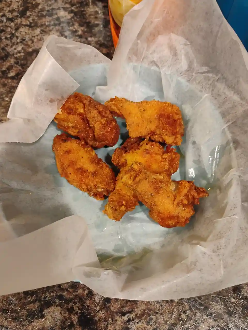 A close-up photo shows a basket lined with white paper, filled with golden-brown, crispy fried chicken wings and pieces. The…