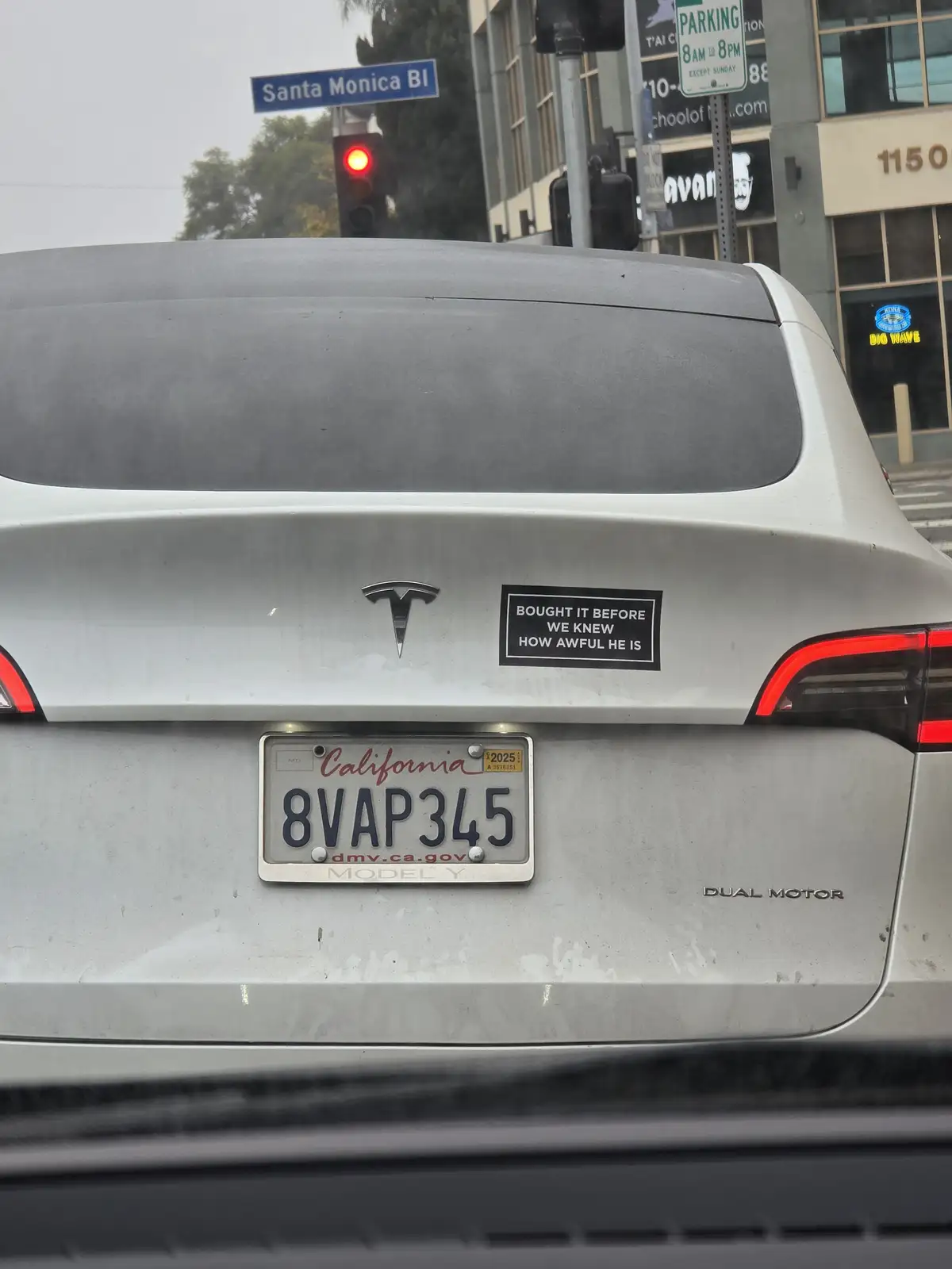 The rear of a silver Tesla Model Y with a California license plate reading '8VAP345' is shown stopped at a red light on…