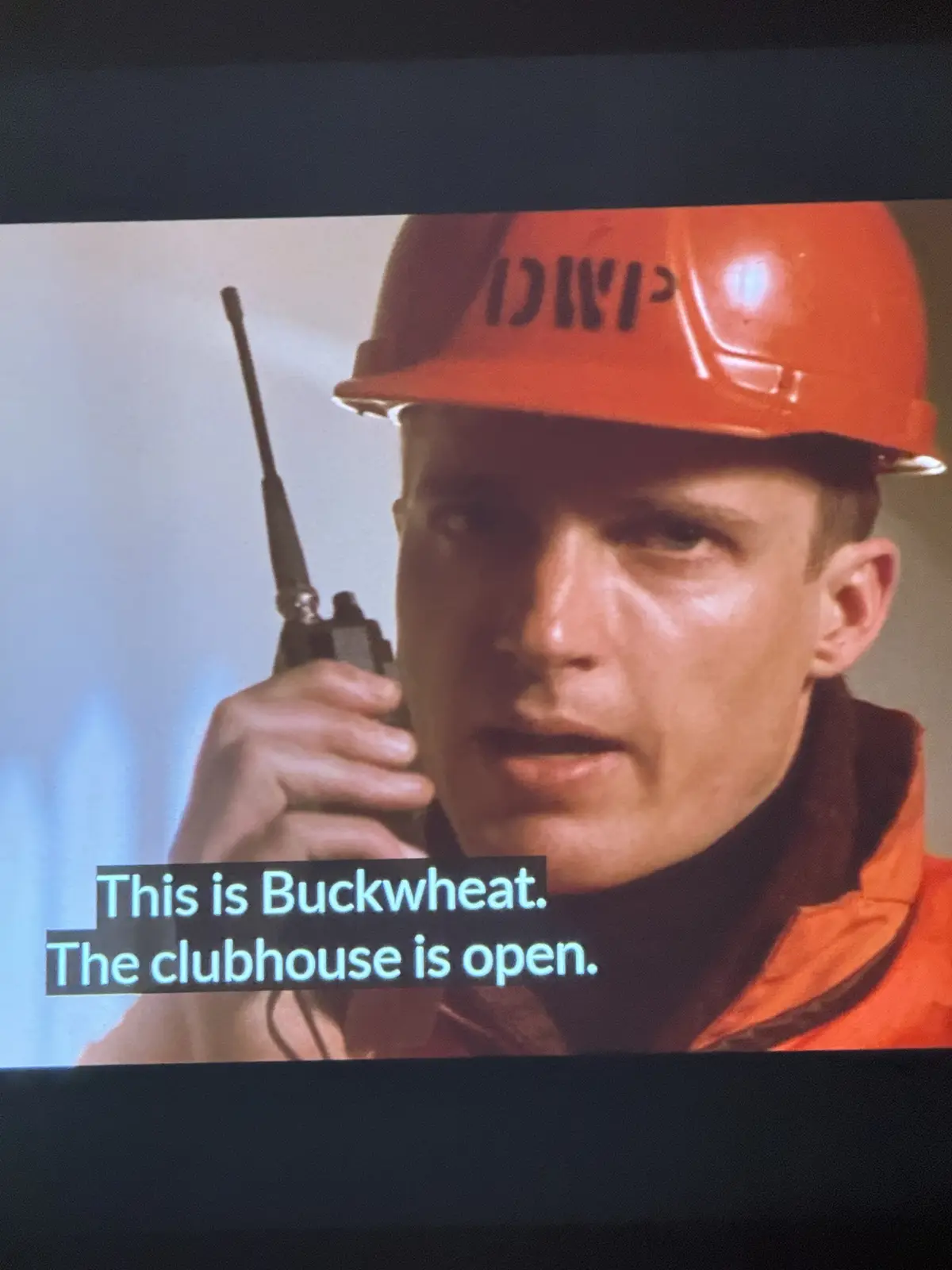 A man wearing an orange hard hat with 'DWP' written on it holds a walkie-talkie to his mouth. The overlaid text reads 'This…