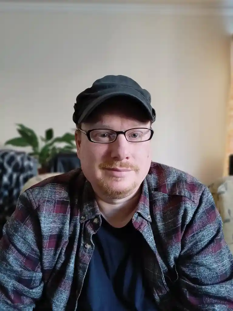 A man with glasses and a black cap looks directly at the camera with a slightly annoyed or unimpressed expression. He wears…