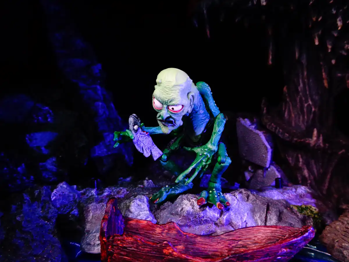 A green, wrinkled, bald monster figurine with glowing red eyes crouches on a rocky surface, holding a small purple fish. The…