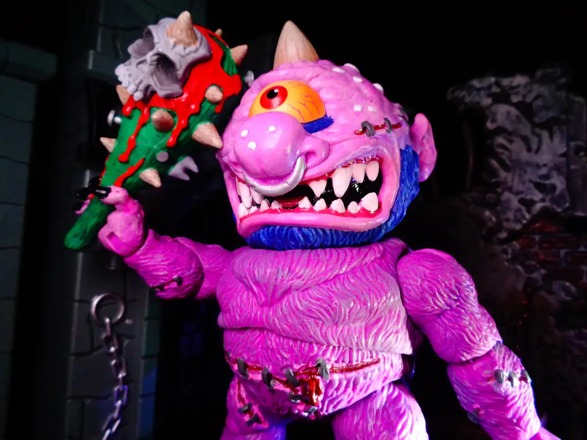 A pink, one-eyed, stitched-up monster toy with a spiked mace topped with a skull, posed aggressively against a dark…