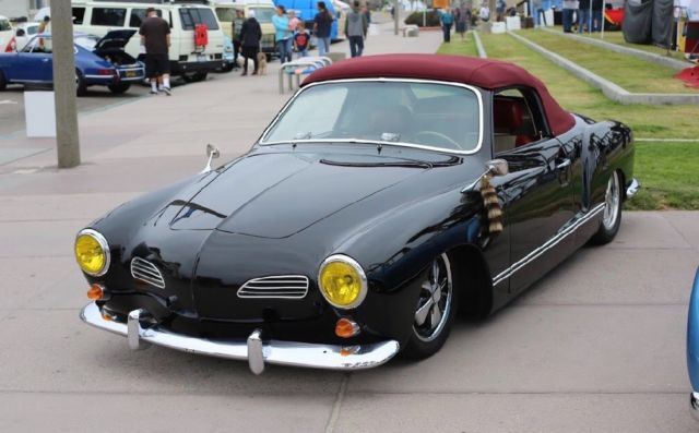 A black vintage Volkswagen Karmann Ghia convertible with a red soft top is parked on a sidewalk at what appears to be a car…