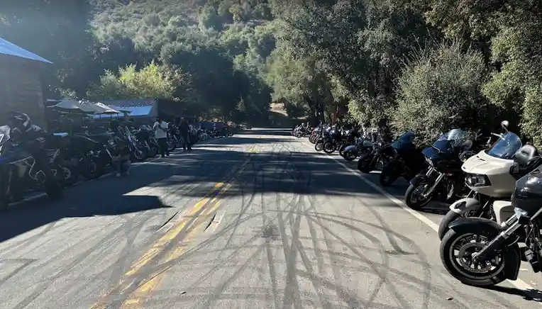 A wide-angle photo of a paved road flanked by rows of parked motorcycles under trees, with people milling about. The road…