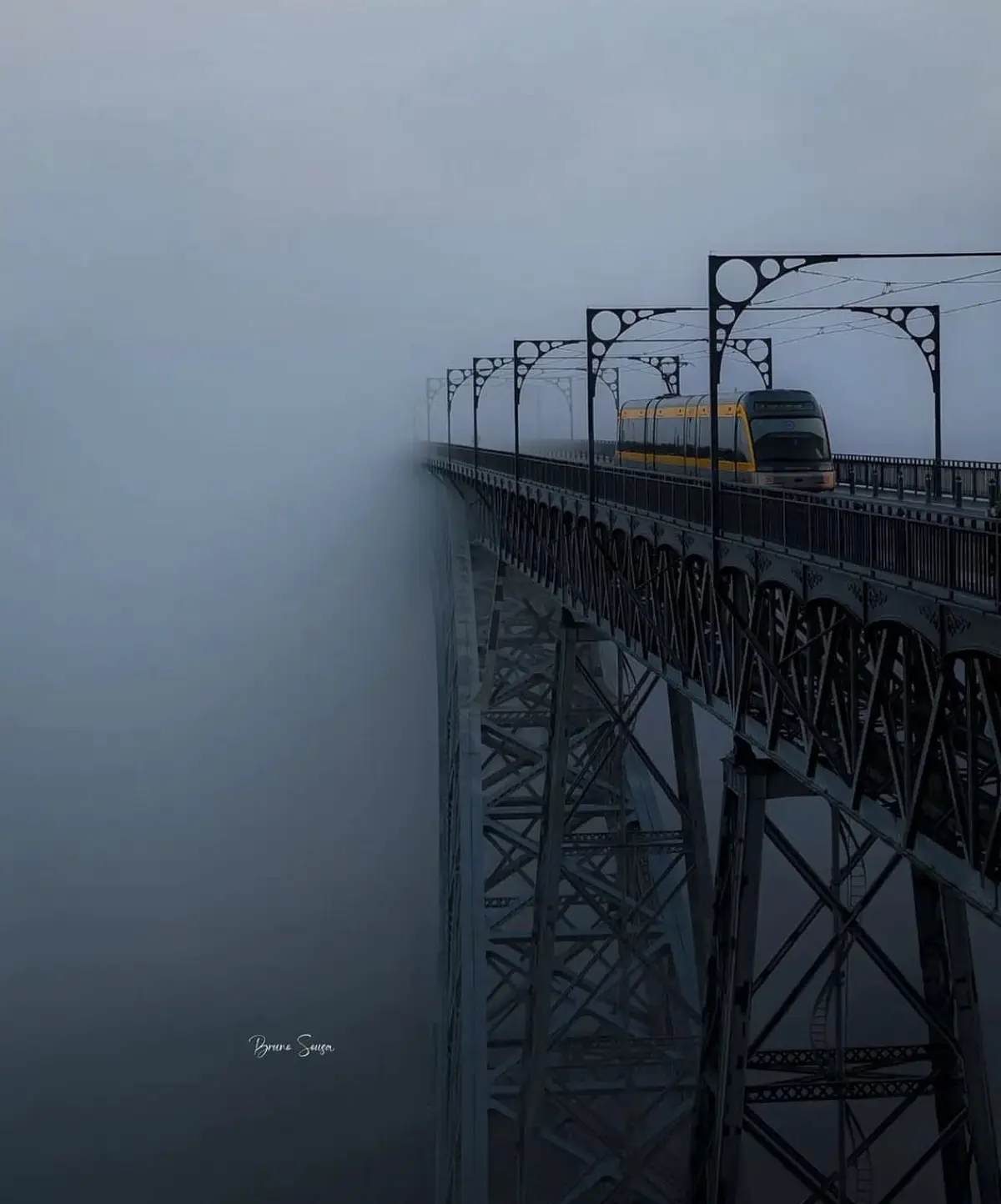 A yellow and grey tram travels across a high, ornate metal bridge enveloped in thick fog, with the bridge's intricate truss…