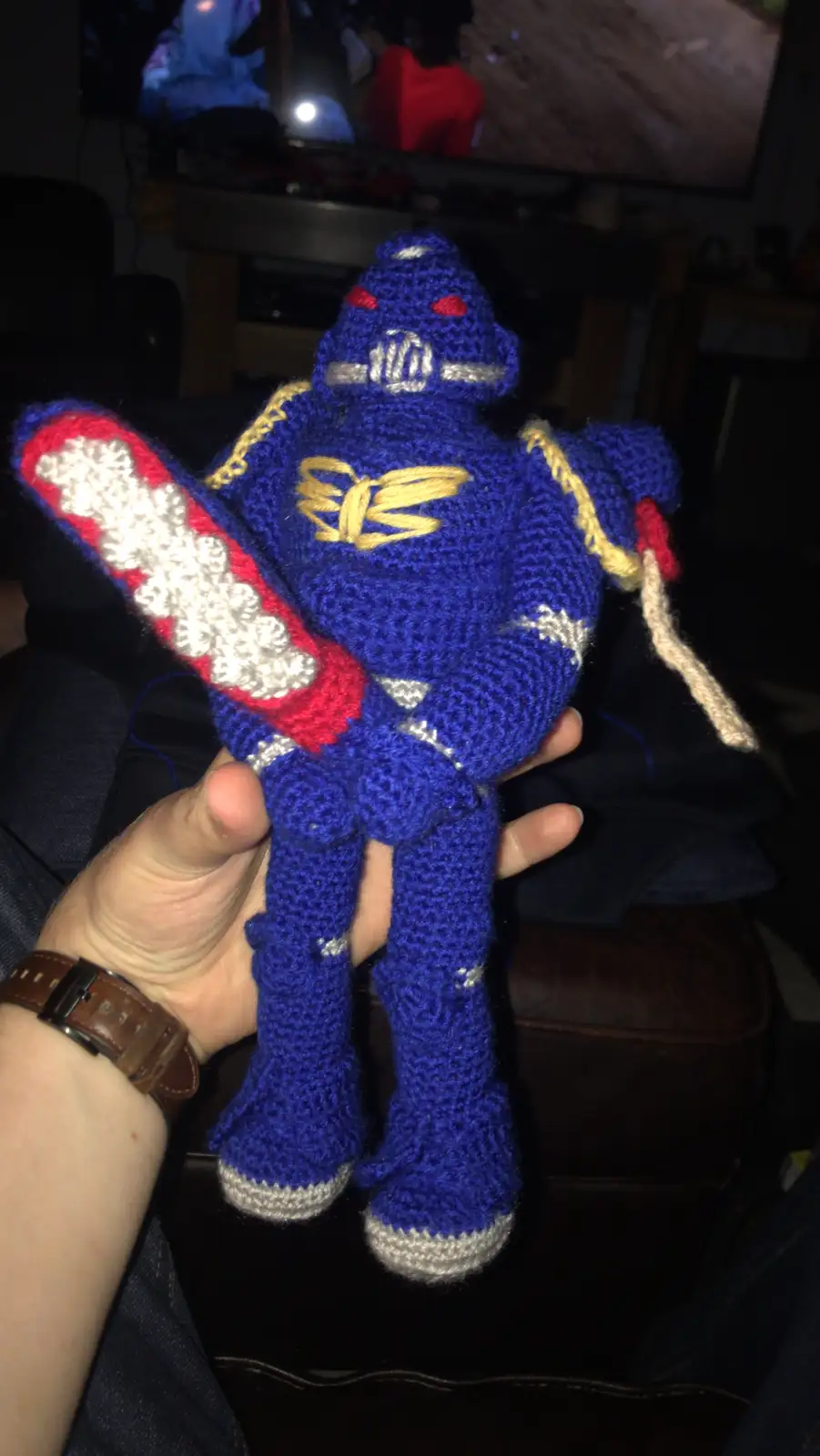 A hand holds a blue crocheted robot toy with red eyes, silver accents, and a red-and-white sword. The robot has a…