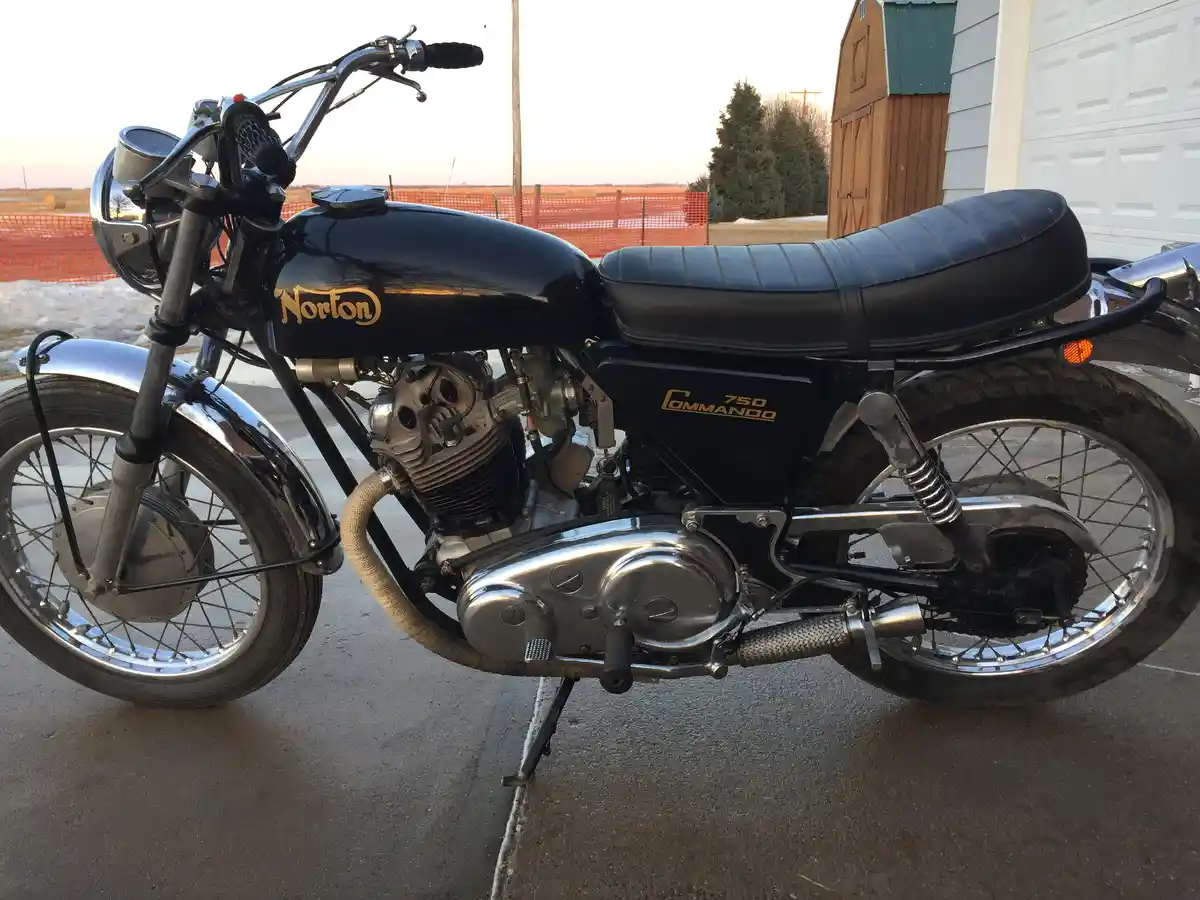 A black Norton 750 Commando motorcycle is parked on a concrete driveway, with its chrome engine and exhaust gleaming. The…