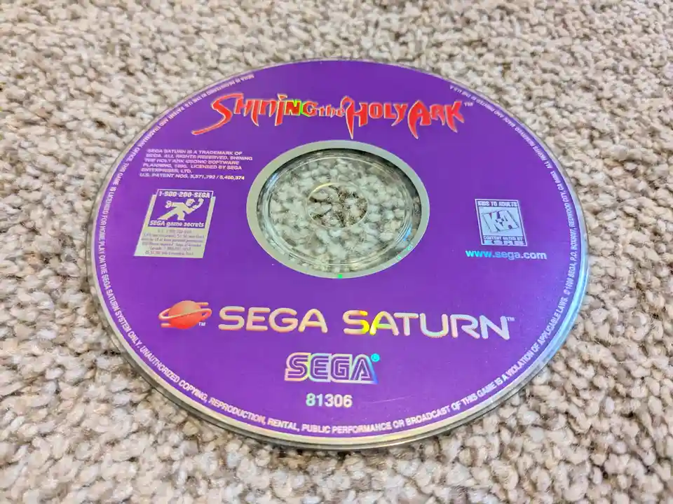 A purple Sega Saturn game disc for 'Shining the Holy Ark' lies on a beige carpet, its central hole filled with visible white…