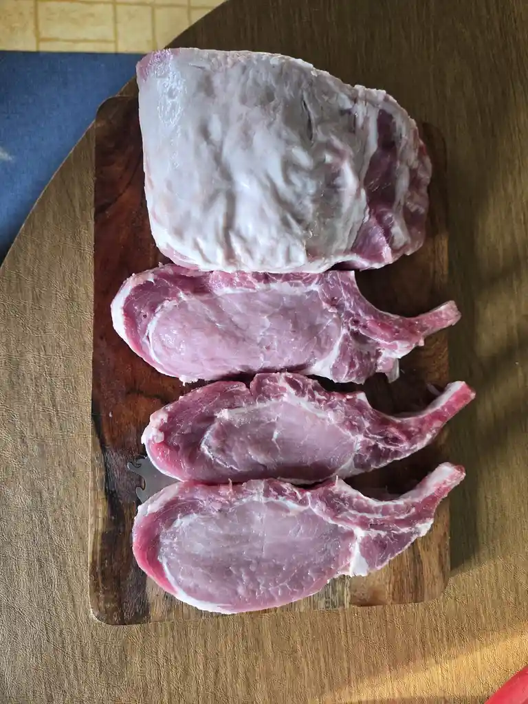 A top-down photo shows four raw pork chops arranged vertically on a dark wooden cutting board, with a larger piece of pork…