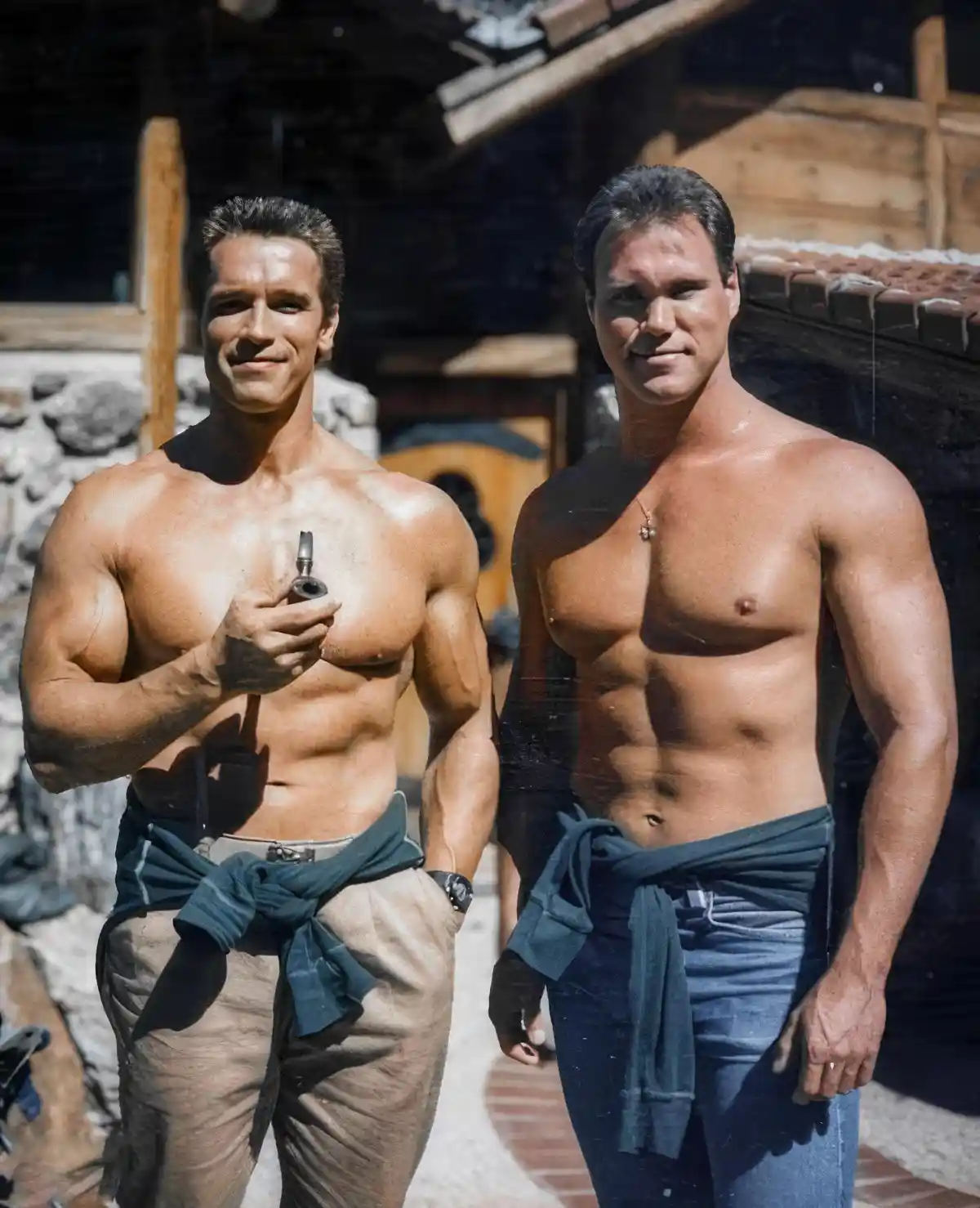Two shirtless, muscular men, identified as Arnold Schwarzenegger and Dorian Yates, stand side-by-side outdoors near a rustic…