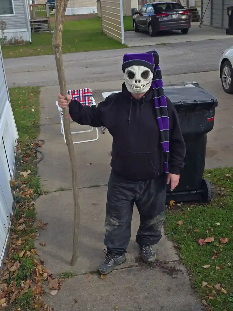 A person wearing a skull mask, a purple and black striped scarf, and a black hoodie stands on a sidewalk holding a plain…