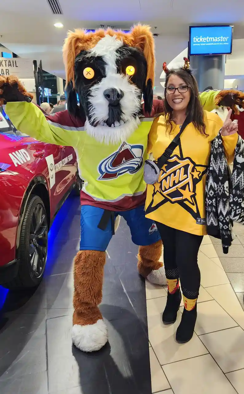 A woman in a yellow NHL jersey poses with a large dog mascot wearing a Colorado Avalanche jersey. The mascot's eyes are…