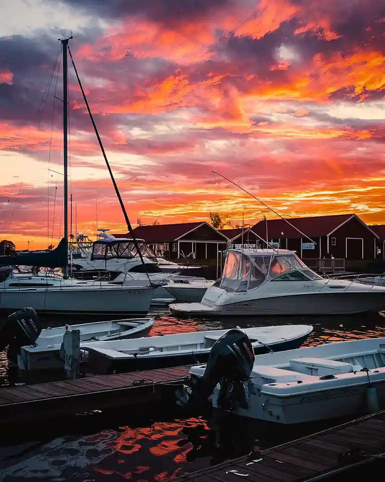 A serene marina at sunset with boats docked, vibrant orange and pink clouds reflecting on the water, and red-roofed…