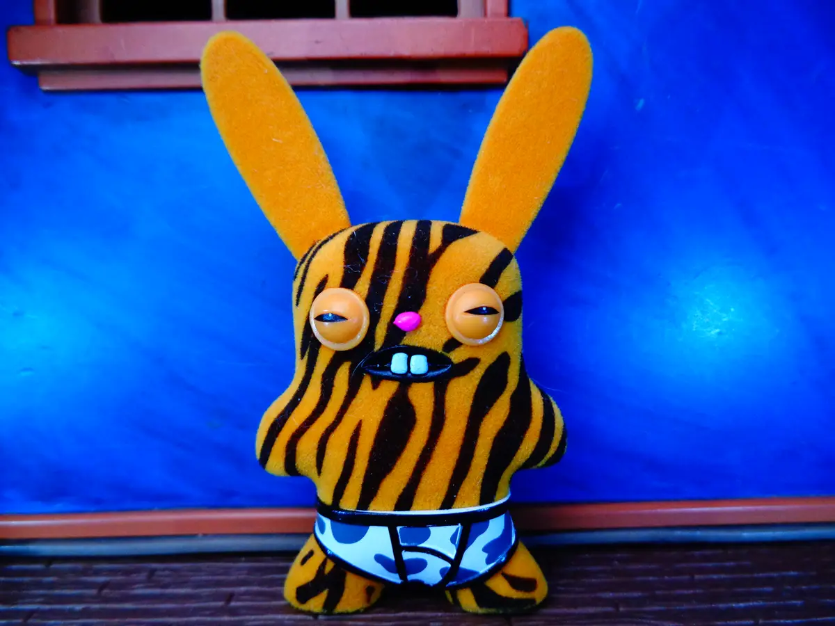 A plush toy resembling a rabbit with orange and black tiger stripes, large round eyes, a pink nose, and two white teeth…