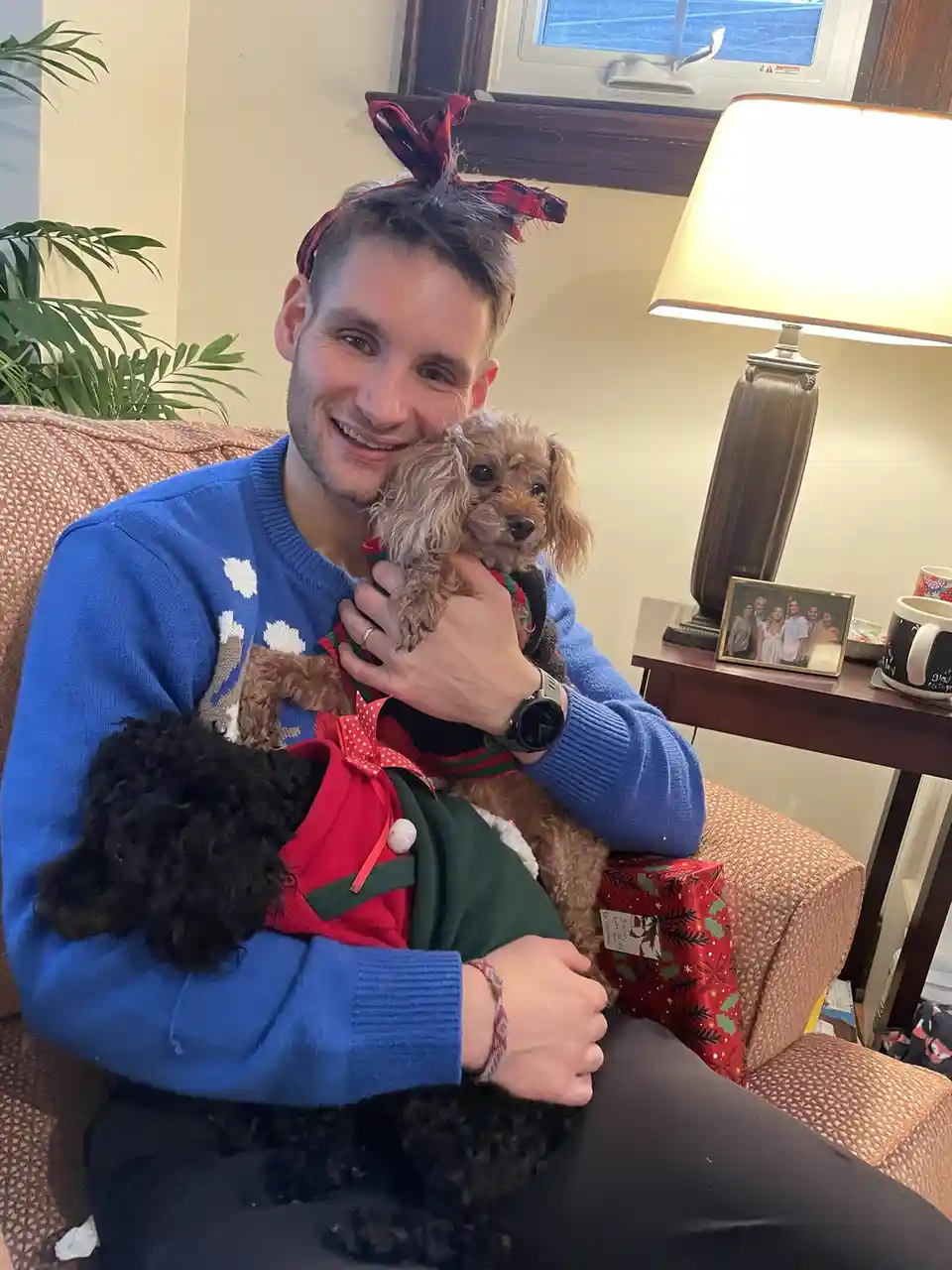 A smiling man wearing a blue holiday sweater and a red plaid bow in his hair sits on a couch holding two small dogs dressed…