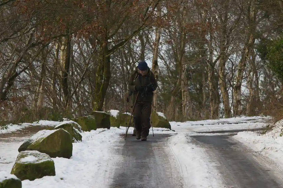 A person in winter gear, including a blue beanie and carrying hiking poles, walks down a snowy, partially cleared path in a…