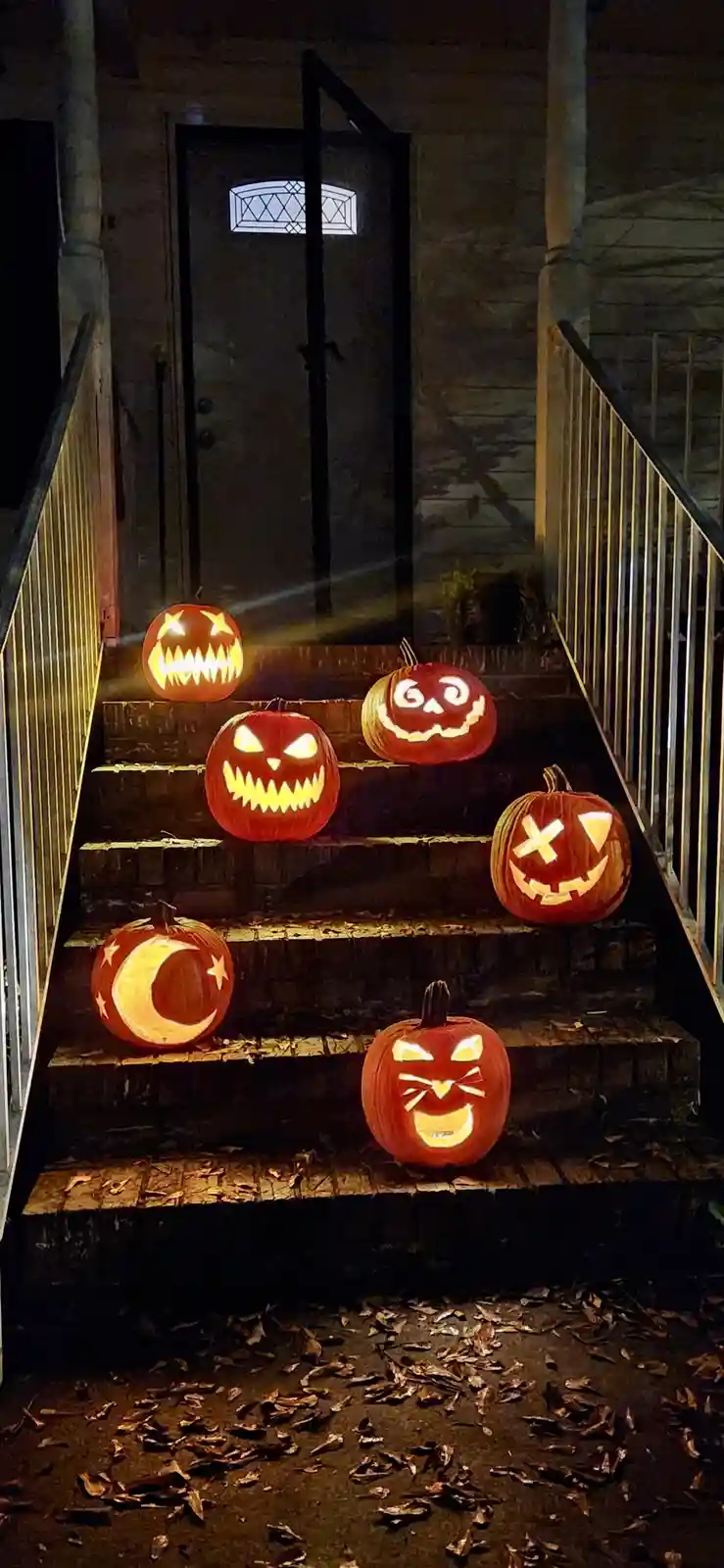 A nighttime photo shows seven glowing jack-o'-lanterns with various carved faces arranged on outdoor stone steps leading to…