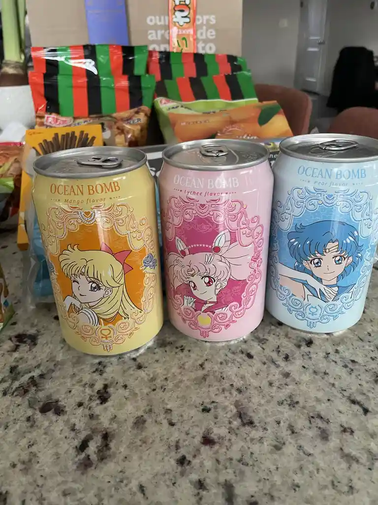 Three colorful 'Ocean Bomb' soda cans featuring anime characters from Sailor Moon are lined up on a speckled countertop. The…