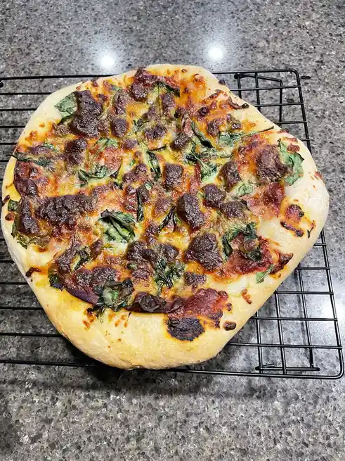A round pizza with a golden-brown crust sits on a black wire cooling rack atop a speckled gray countertop. The cheese is…