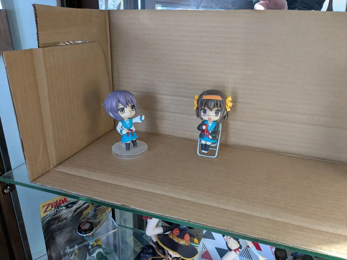 Two chibi anime figurines are positioned inside a cardboard box diorama. One figurine with purple hair stands and gestures…