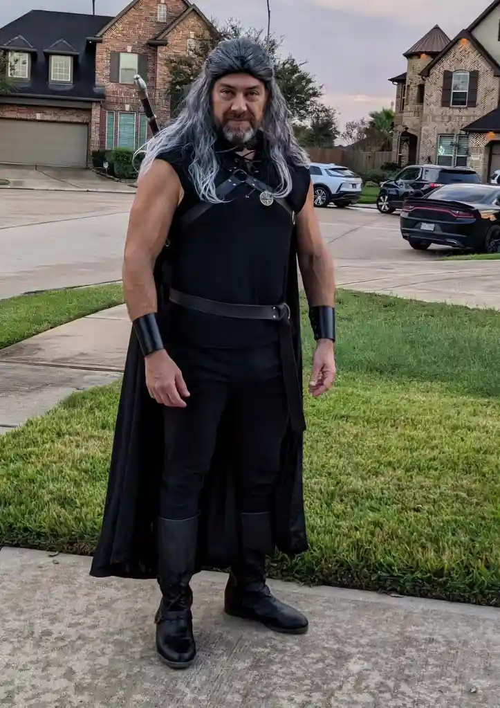 A man in a full black fantasy warrior costume with long grey wig, cape, and sword stands on a suburban sidewalk. The…