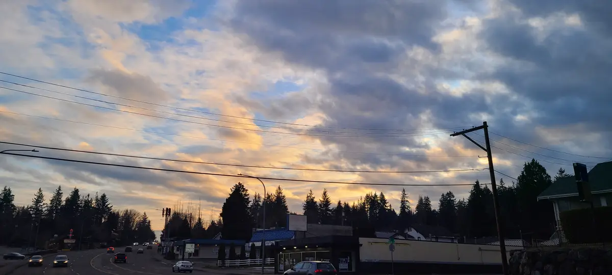 A scenic photo of a suburban street at dusk, with cars on the road, trees in the background, and a dramatic sky with clouds…
