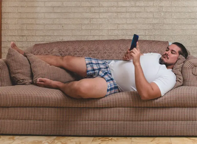 a man with a beard and white t-shirt lies on a brown patterned couch, holding a phone and appearing to be asleep or resting…