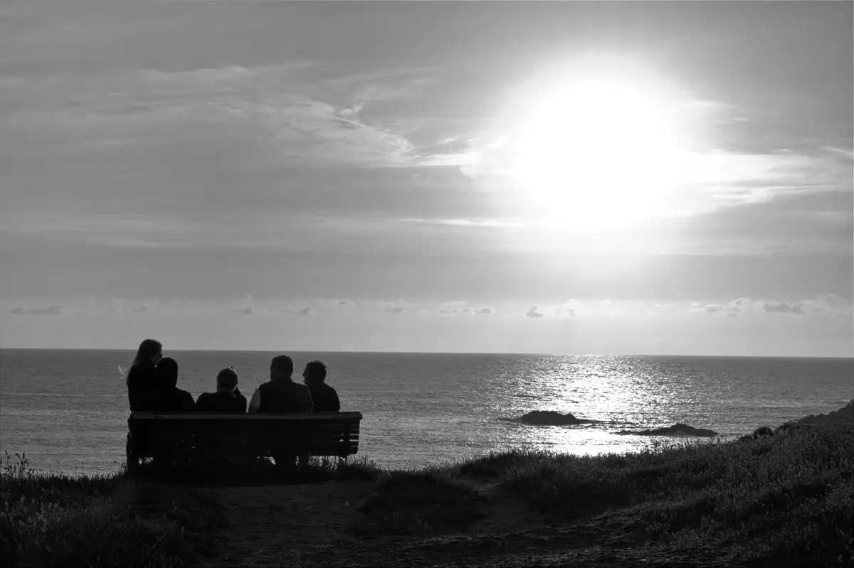 A black-and-white photo shows five people sitting on a bench overlooking the ocean, silhouetted against a bright sun and…