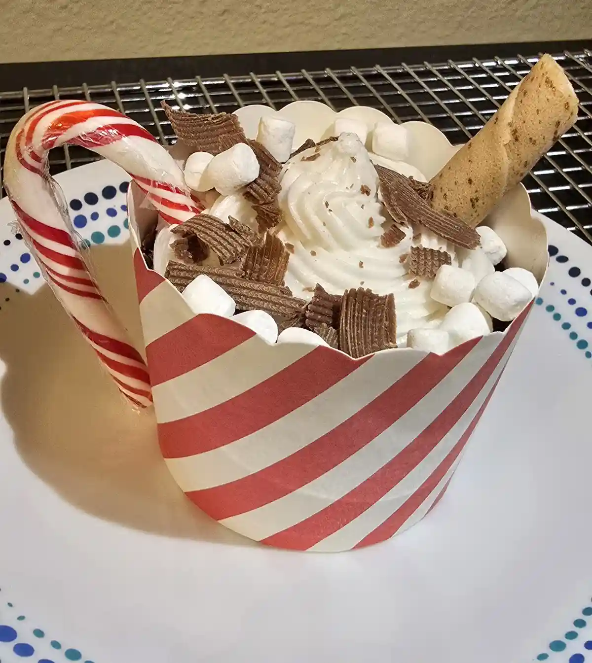 A red-and-white striped paper cup holds a dessert topped with whipped cream, mini marshmallows, chocolate wafer pieces, and…