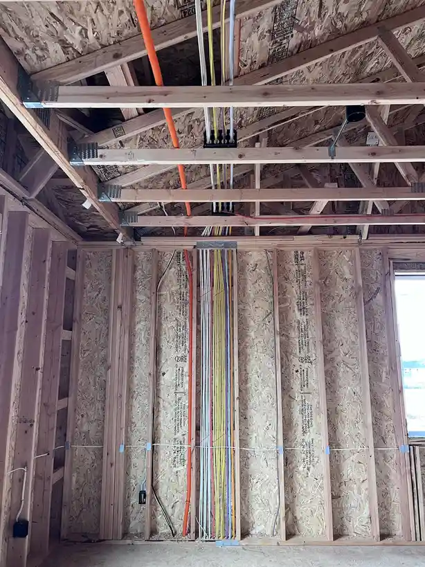 An image of a construction site showing exposed wooden framing, OSB sheathing, and a large bundle of colorful electrical…