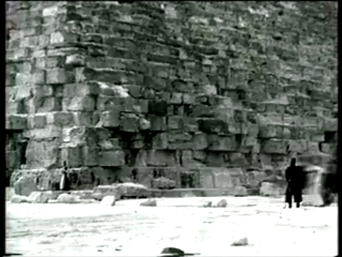 A grainy black-and-white historical photo shows a lone, silhouetted figure standing before a massive, ancient stone wall…