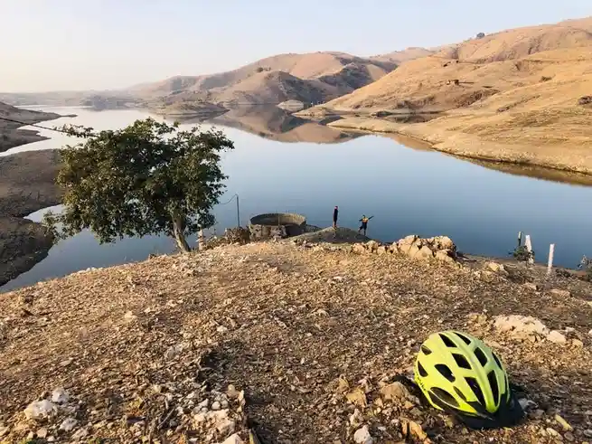 A bright yellow bicycle helmet rests on dry, rocky ground in the foreground, overlooking a calm lake surrounded by arid…