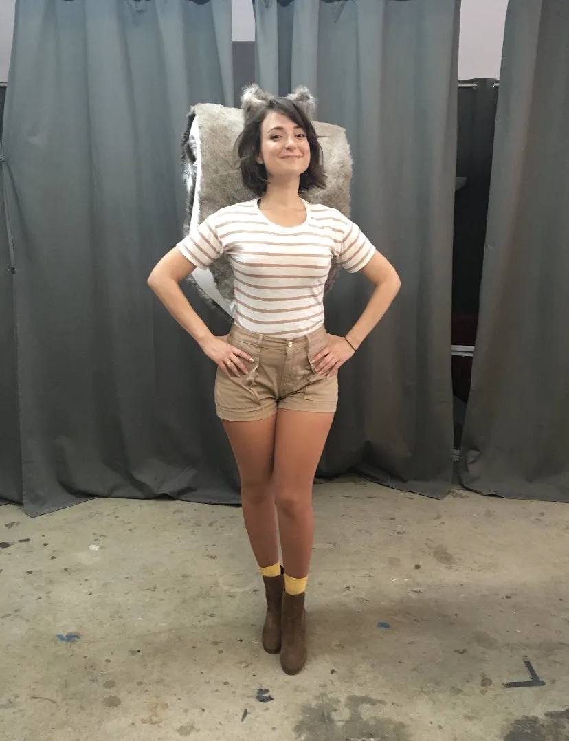 A woman stands confidently with hands on hips, wearing a striped shirt, shorts, and boots, with small wolf ears on her head…