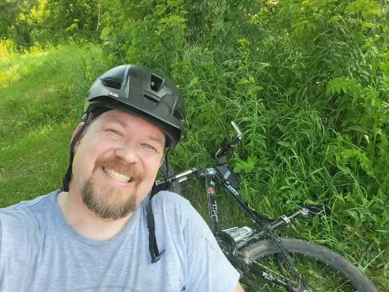 A man wearing a black bicycle helmet and a light blue t-shirt smiles broadly at the camera while lying in a grassy field…