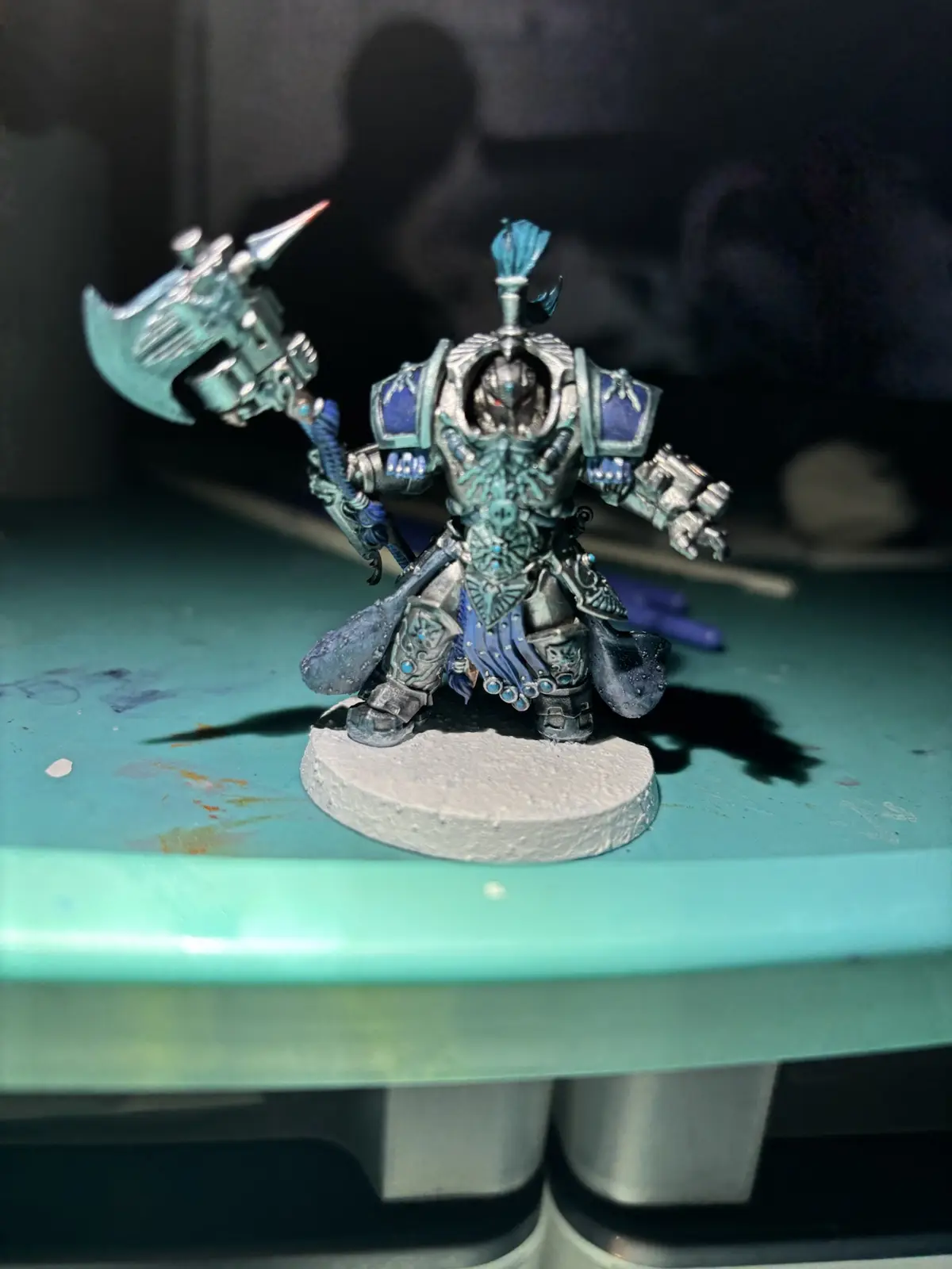 A heavily armored miniature warrior figure, painted in shades of teal and blue with silver accents, stands on a circular…