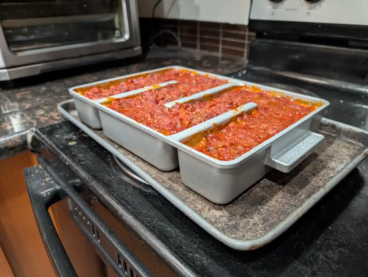 Three small, identical metal baking pans filled with red tomato sauce sit on a larger metal baking sheet atop a black…