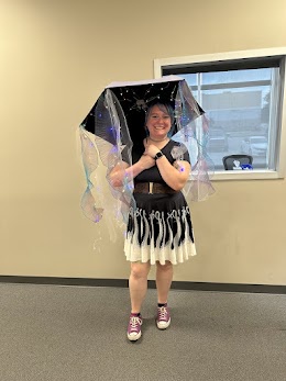 A person in a black and white costume with translucent fairy wings and a starry umbrella stands indoors smiling. The text…