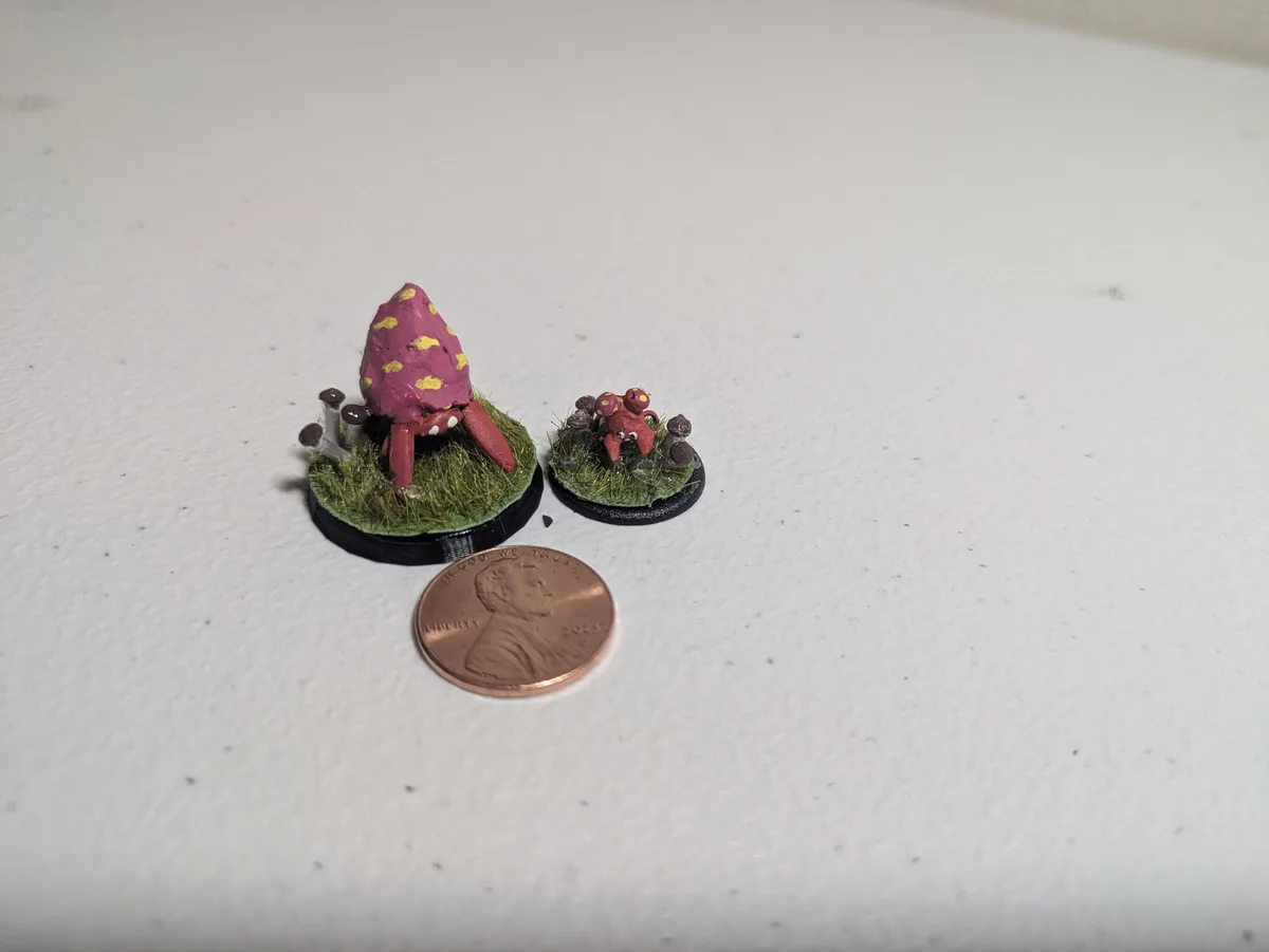 Two tiny hand-painted miniature crab-like creatures with shells stand on grassy bases next to a U.S. penny for scale. One…
