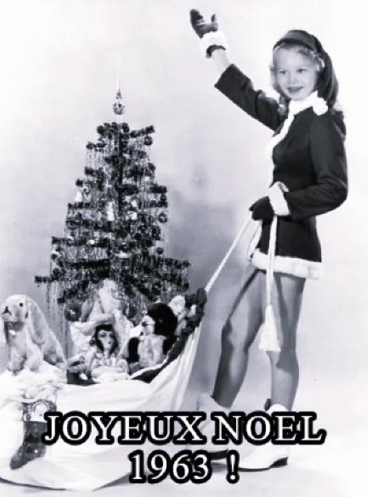 A vintage black-and-white photo shows a young girl in a festive outfit standing beside a decorated Christmas tree, holding a…