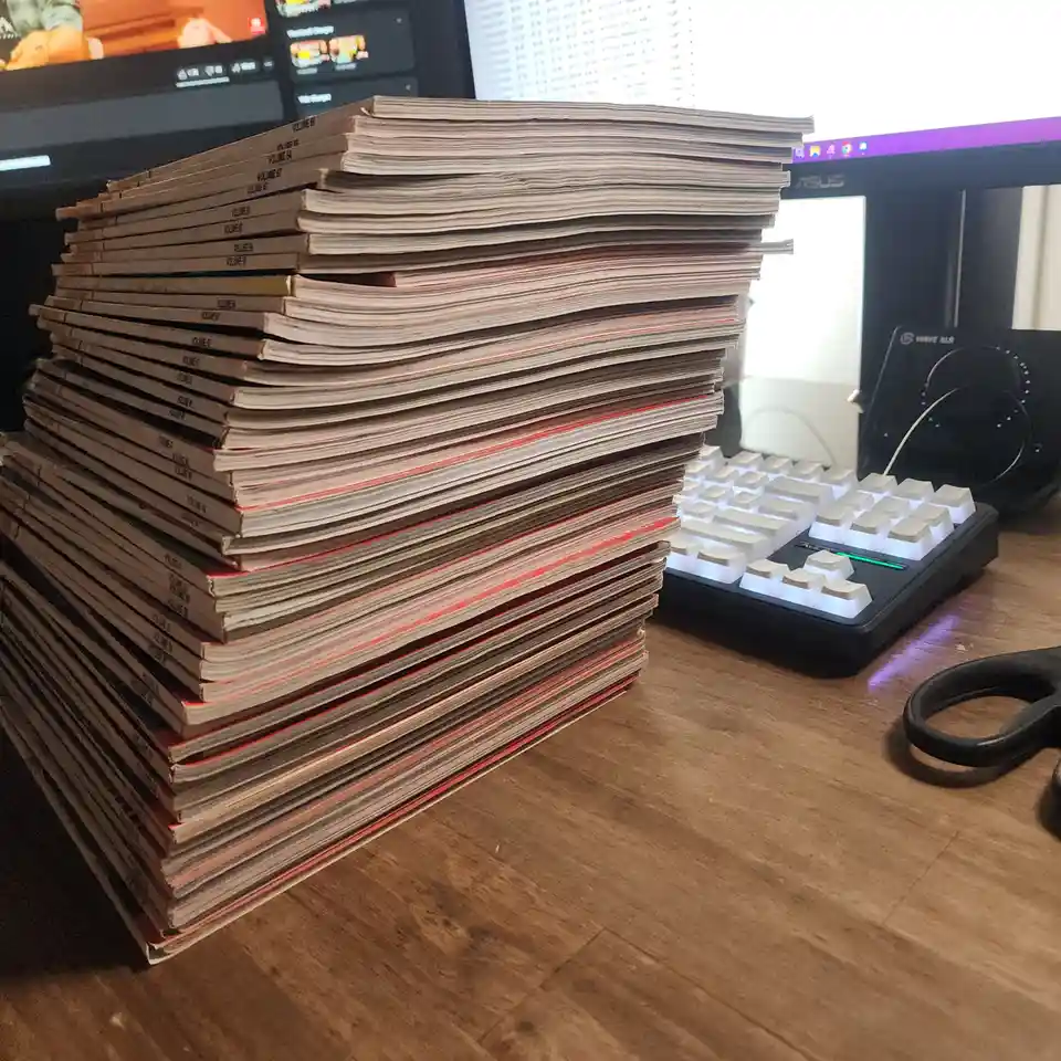 A towering stack of manga volumes sits on a wooden desk next to a white mechanical keyboard, a pair of black scissors, and…