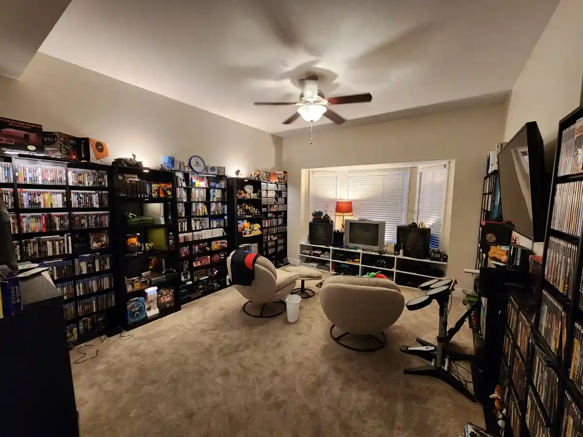 A wide-angle photo of a cozy, carpeted room filled with black shelving units packed with video games, consoles, and…
