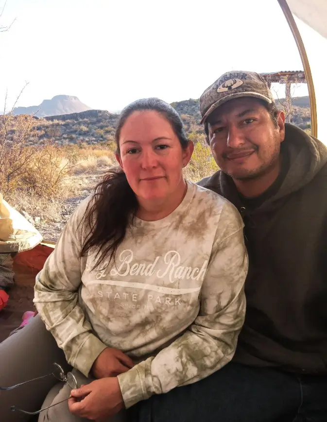 A man and woman are sitting together inside a tent, smiling at the camera with a desert landscape and a distant mountain…