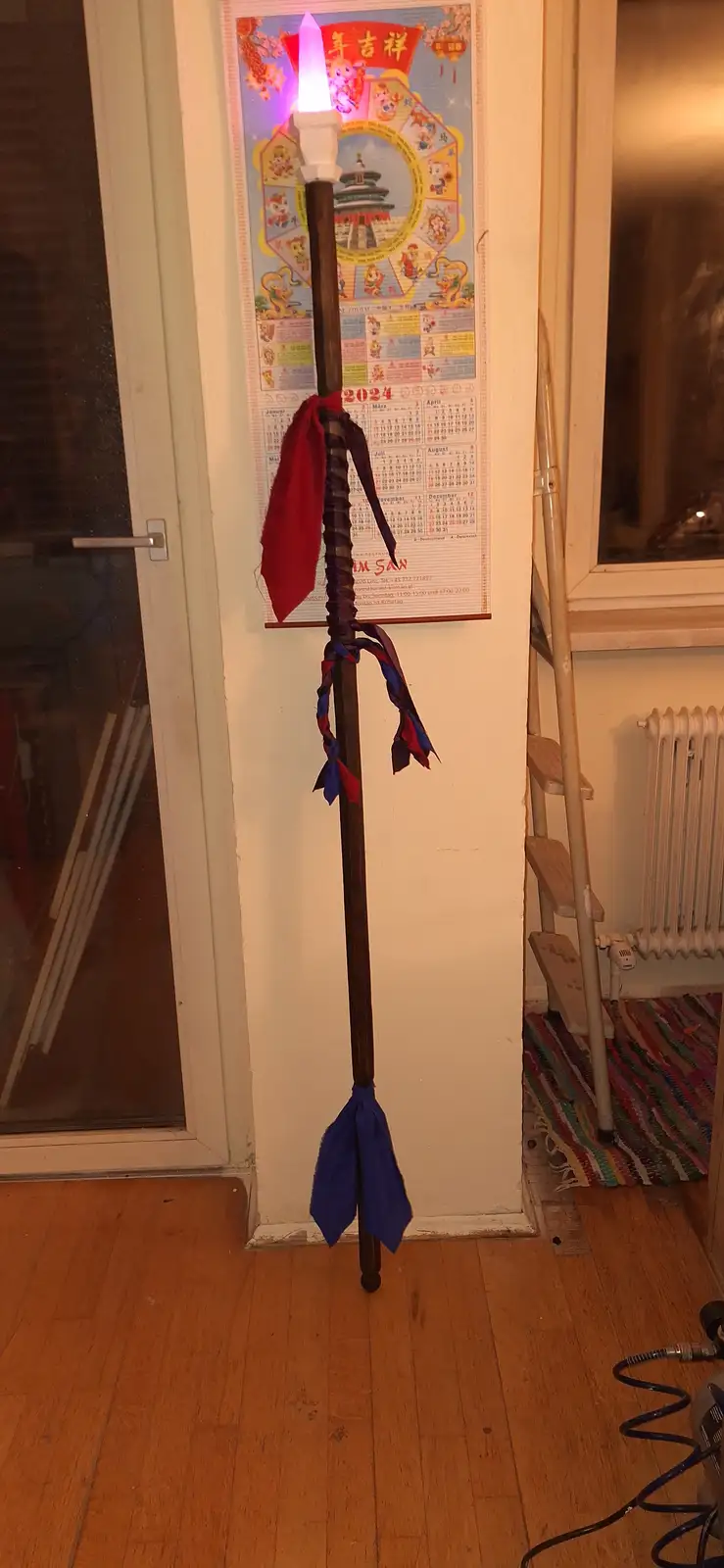 A tall wooden staff decorated with red and blue ribbons stands upright in a room, topped with a glowing purple LED light…