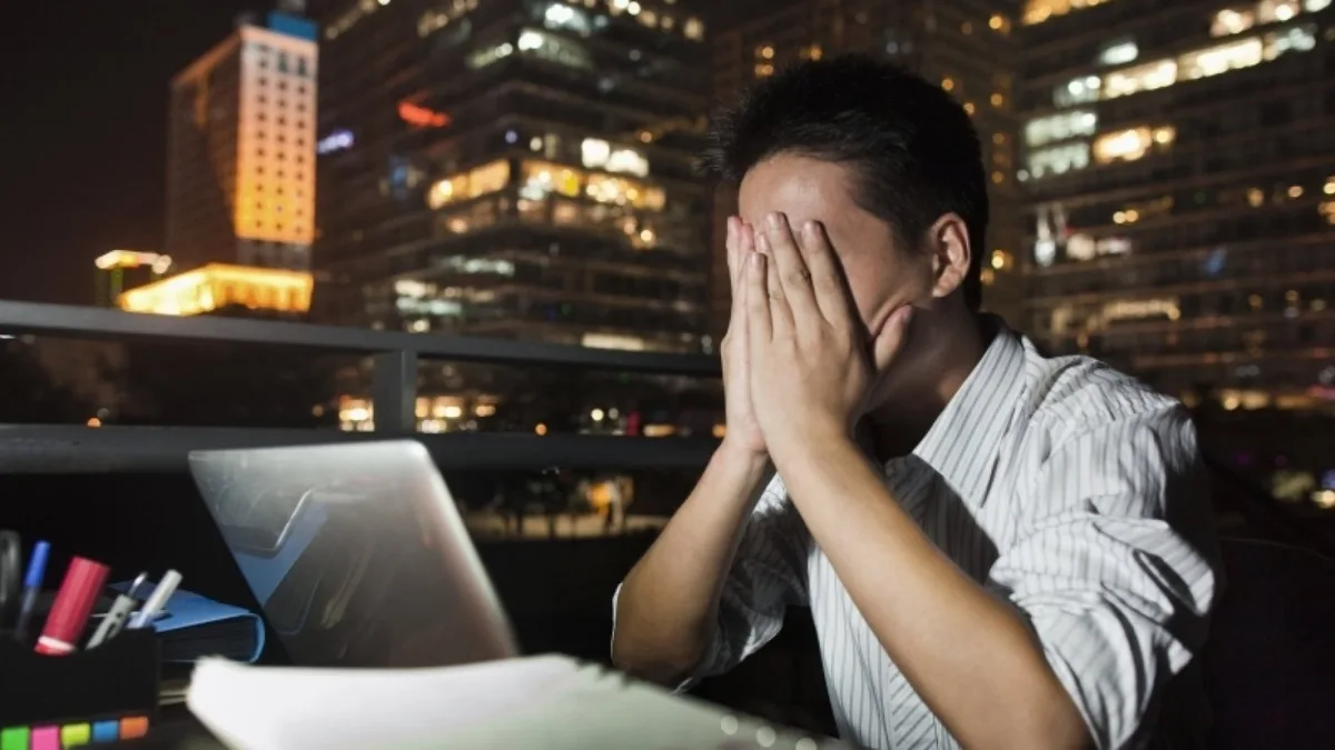 A man in a striped shirt sits at a desk with a laptop and papers, covering his face with both hands in exhaustion. Behind…