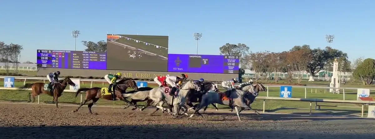 A wide shot of a horse race in progress at Fair Grounds, with jockeys on horses galloping on a dirt track. A large digital…