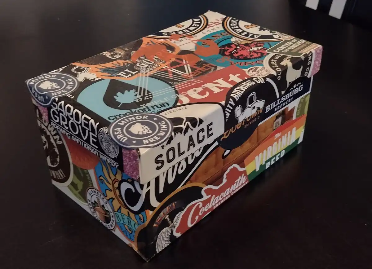A rectangular cardboard box covered in a collage of colorful beer brewery stickers from various Virginia breweries…