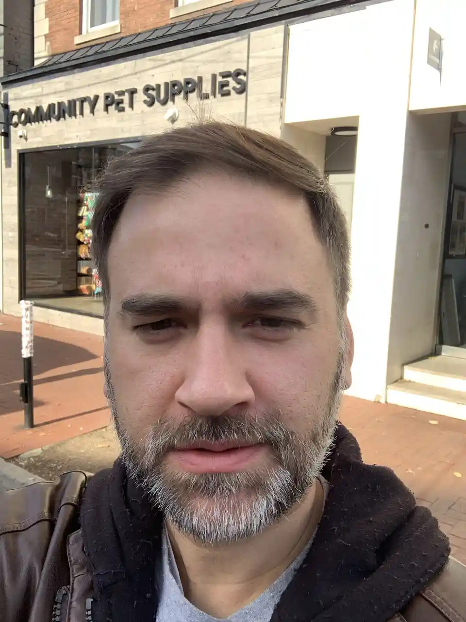 A man with a graying beard and tired expression takes a selfie in front of a store named 'COMMUNITY PET SUPPLIES'. The text…