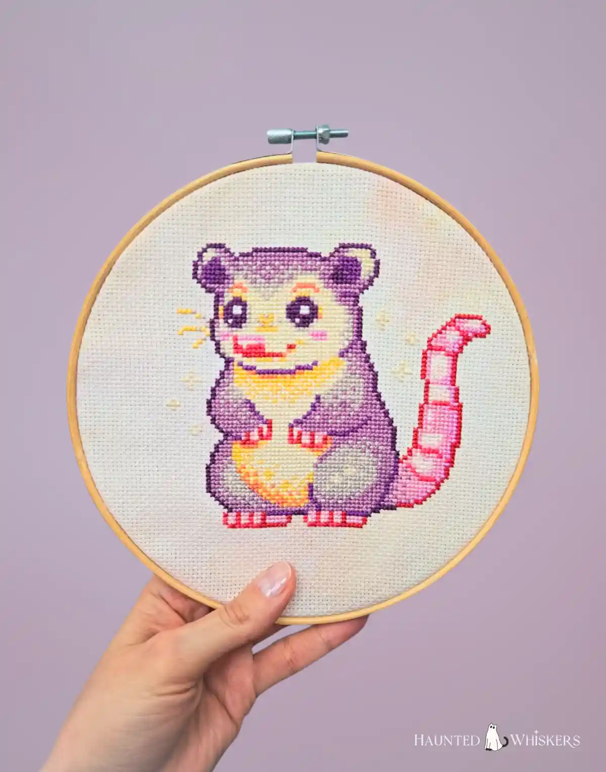 A hand holds a wooden embroidery hoop displaying a colorful cross-stitch of a cute, cartoonish purple opossum with a pink…