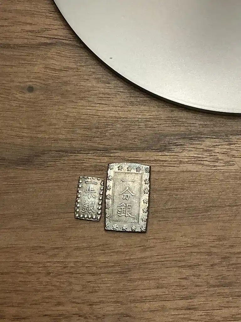 Two small, rectangular silver-colored Japanese coins labeled '一分銀' and '一朱銀' lie on a wooden surface next to a white plate…